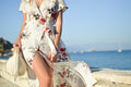 Woman in a breezy linen wrap midi dress walking along a sunlit seaside path with a woven bag — placeholder image
